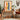 May include: A framed print featuring the word "CHAOS" in a colourful, abstract font. The print is hanging on a wall with a macrame wall hanging and a wooden chair with an orange cushion. There are plants in pots on a wooden bench in front of the print.